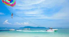Person parasailing above the clear blue water in Phuket, with boats in the background.