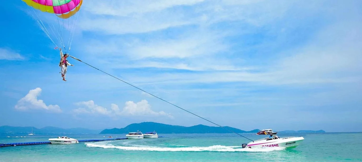 Person parasailing above the clear blue water in Phuket, with boats in the background.