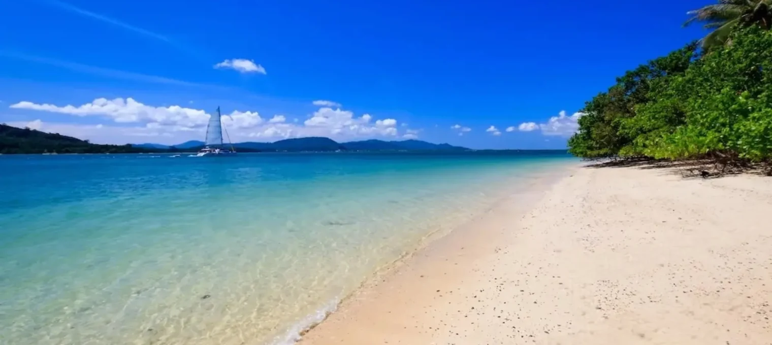 Pristine beach in Phuket with clear water, gentle waves, and a distant sailboat.