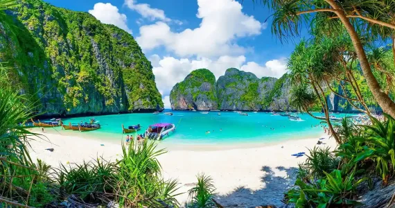 Tropical beach in Phuket with clear water, boats, lush greenery, and dramatic cliffs.