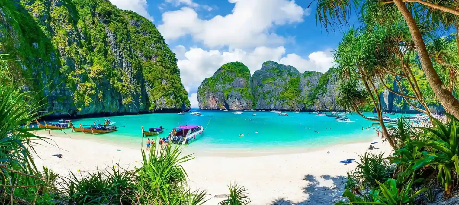 Tropical beach in Phuket with clear water, boats, lush greenery, and dramatic cliffs.