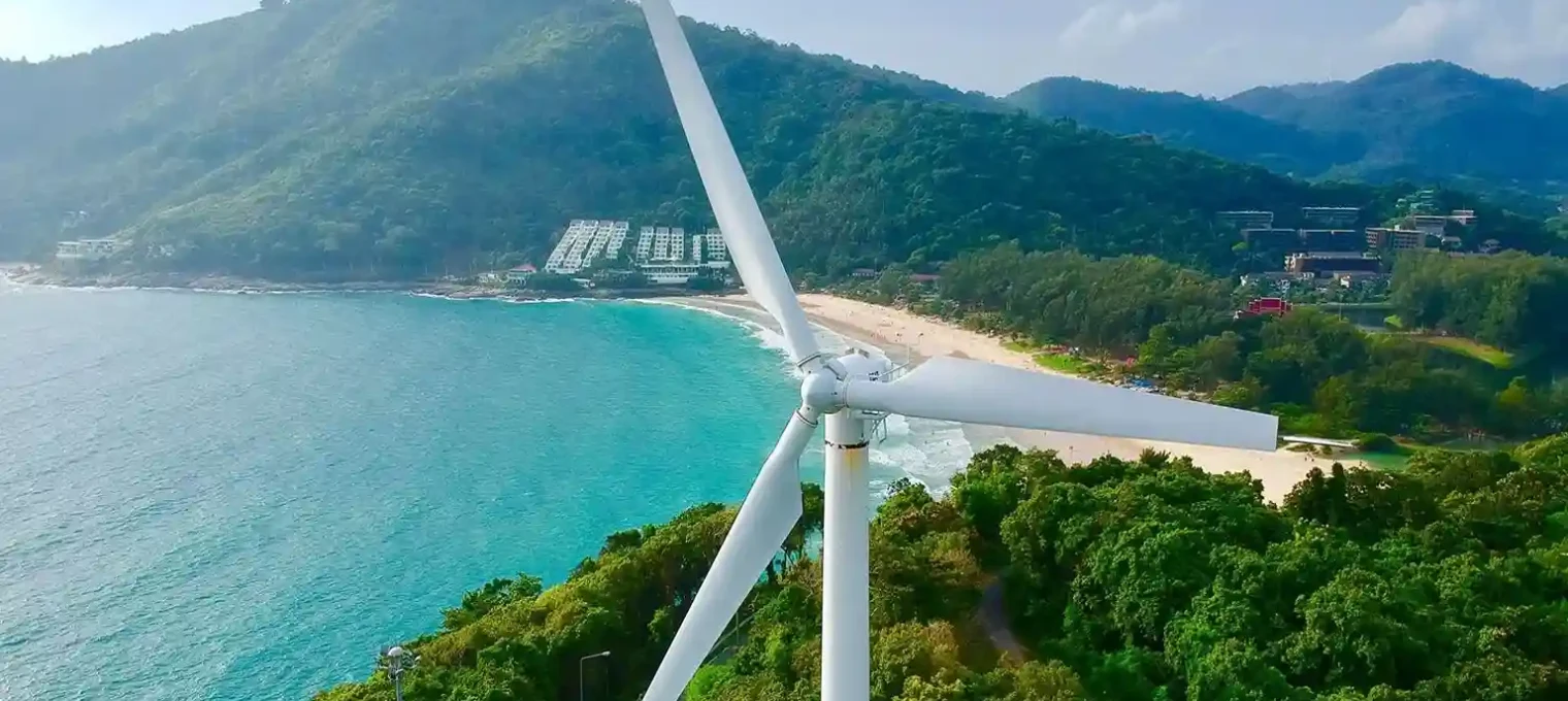Aerial view of a wind turbine overlooking a beach and lush green hills in Phuket.